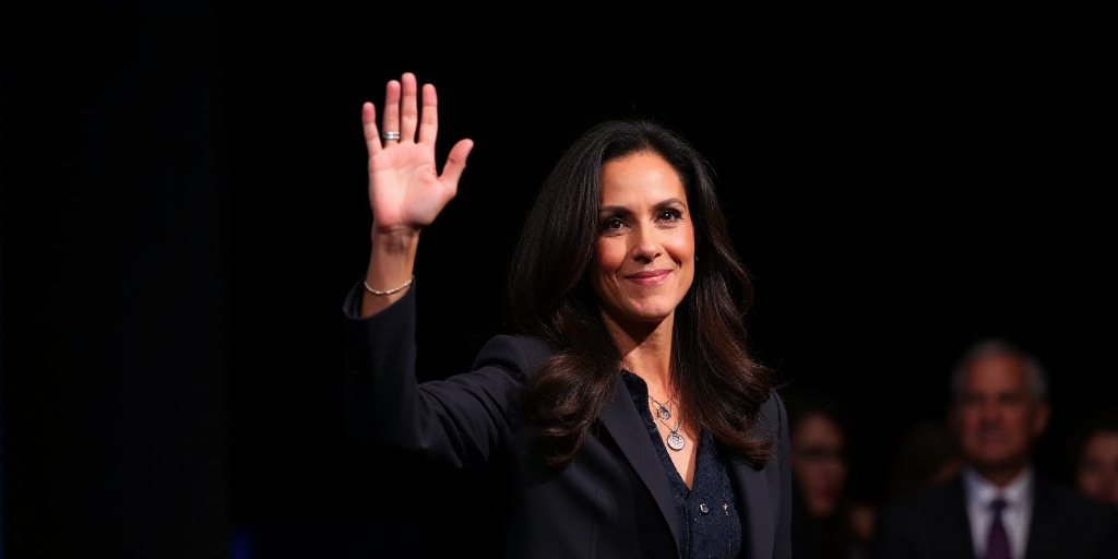 a woman in a suit and tie waves to the crowd at a political event in a dark room with a black backgr