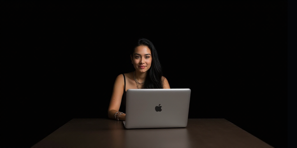a woman sitting at a table with a laptop computer in front of her and a black background behind her,