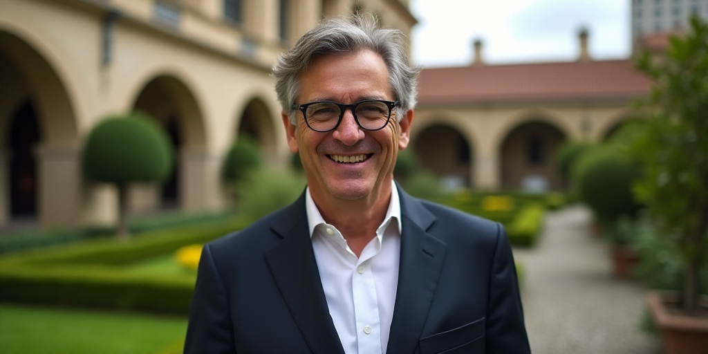 a man in a suit and glasses smiling for the camera with a garden in the background and a building in