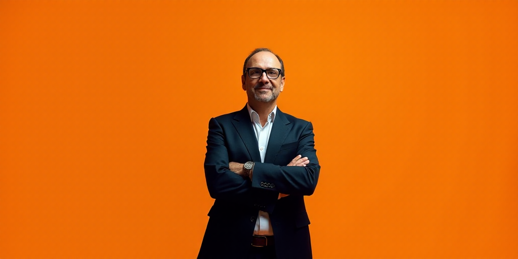 a man in a suit and glasses standing with his arms crossed in front of an orange background with the